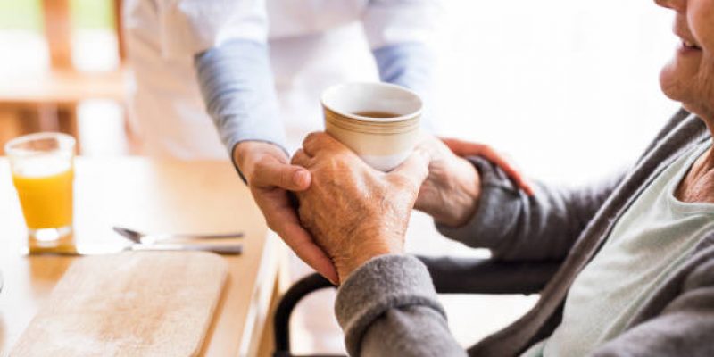 Unrecognizable health visitor and a senior woman during home visit. A nurse giving tea to an elderly woman sitting at the table. Close up.