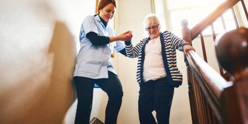 Young female caregiver helping senior woman walking down stairs at home