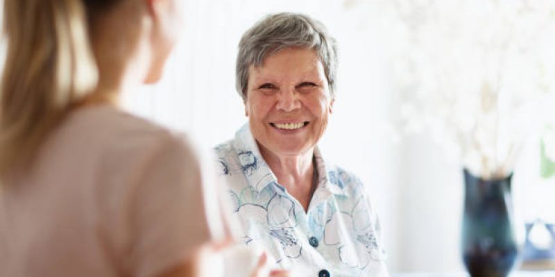 young woman chatting with senior woman, indoors in living room, sitting on sofa. They are having a cup of coffee, smiling at each other